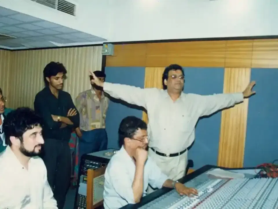 A man with arms outstretched beside a mixing console in a recording studio